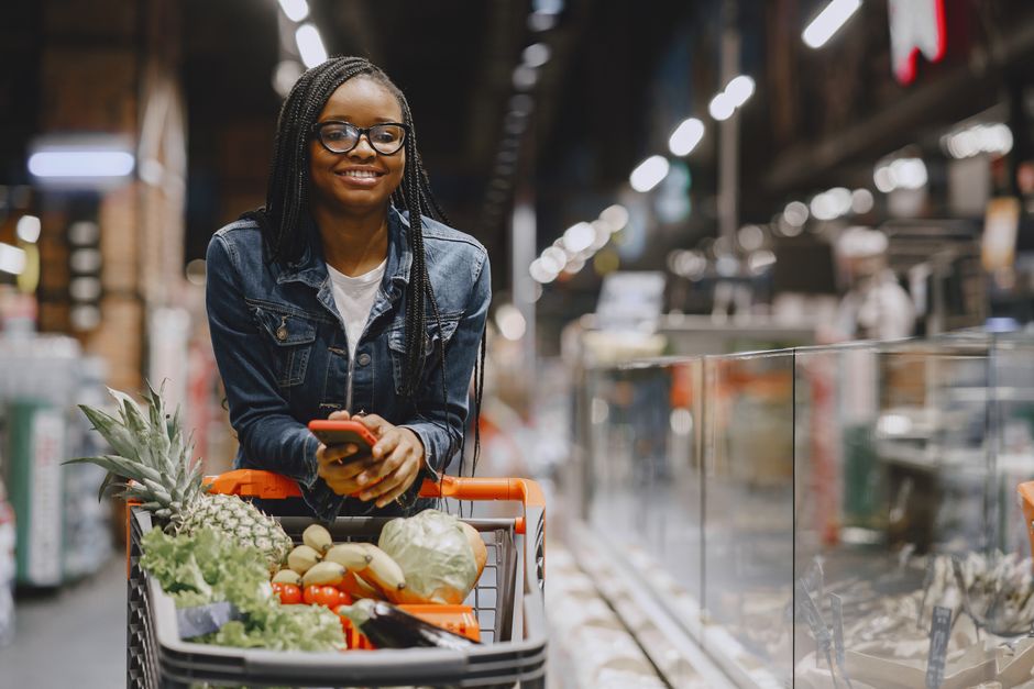 A woman smiles while shopping for fresh produce in a supermarket, using a smartphone.